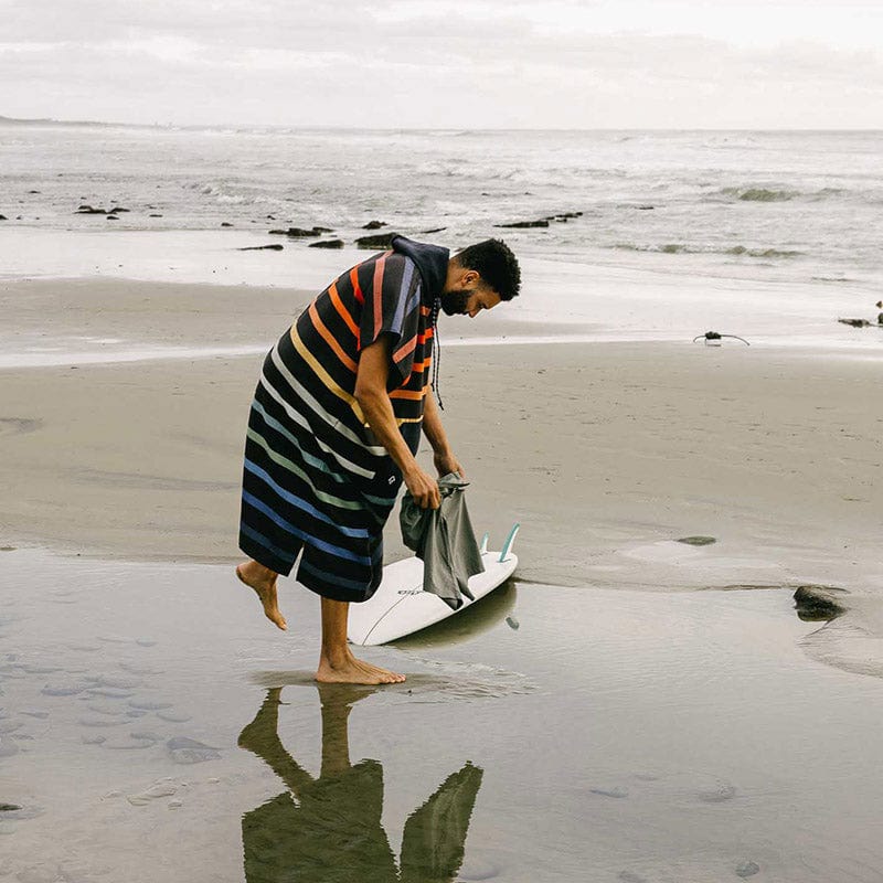 Load image into Gallery viewer, Man on a beach with a striped towel and surfboard, looking at something on the sand.
