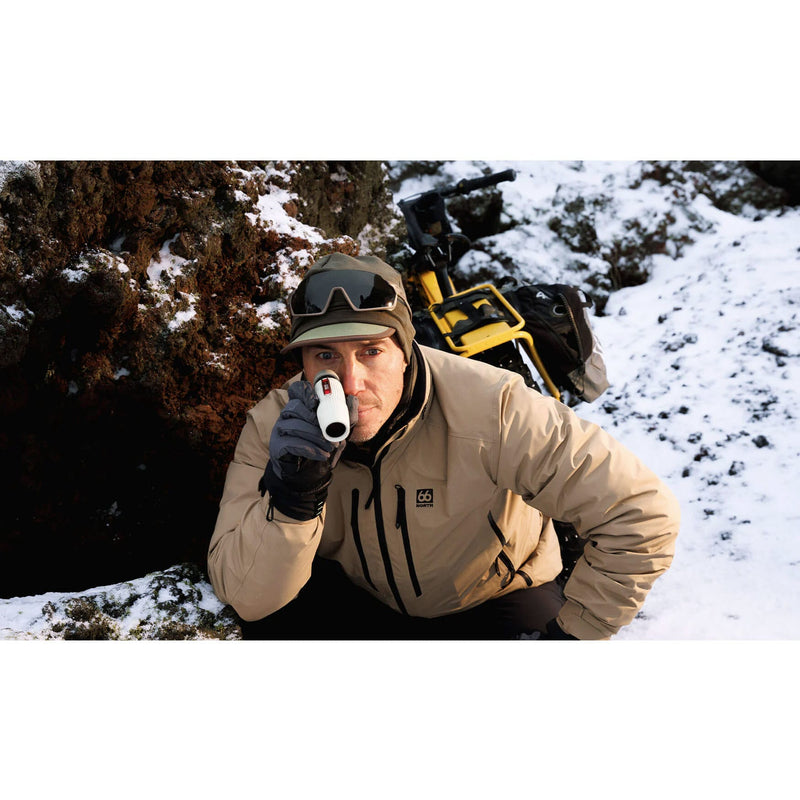 Load image into Gallery viewer, A person in a snowy outdoor setting wearing a beige technical winter jacket, black gloves, and a cap, holding a white cylindrical product to their mouth. A yellow and black backpack is visible behind them.
