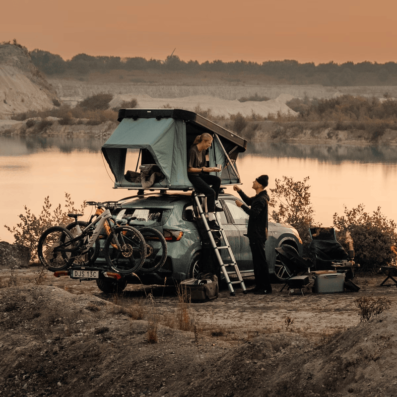 Load image into Gallery viewer, Two people setting up a rooftop tent on a car by a lake at sunset.