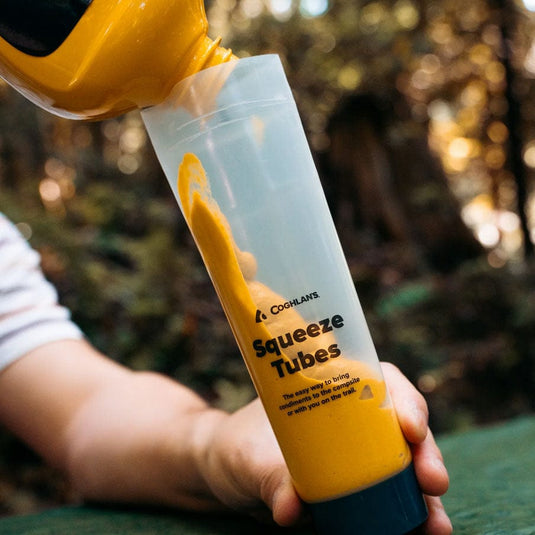 Hand filling a Coghlan's squeeze tube with yellow paint against a blurred natural background