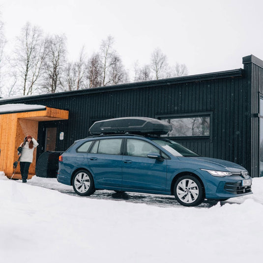Blue car with a roof box parked in front of a modern building in a snowy landscape.