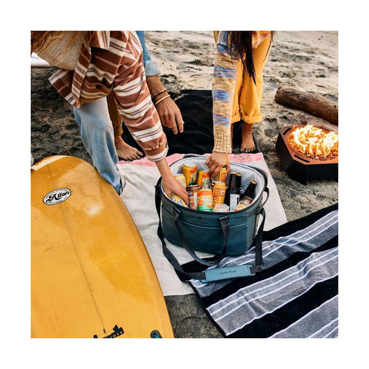Two people by a beach with a cooler full of drinks, a surfboard, and a fire pit.