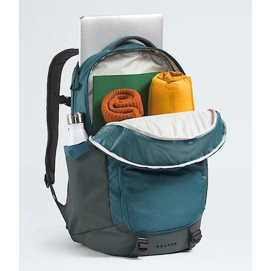 Teal and gray backpack with items including a laptop, green book, orange towel, and yellow bag on a light gray background.