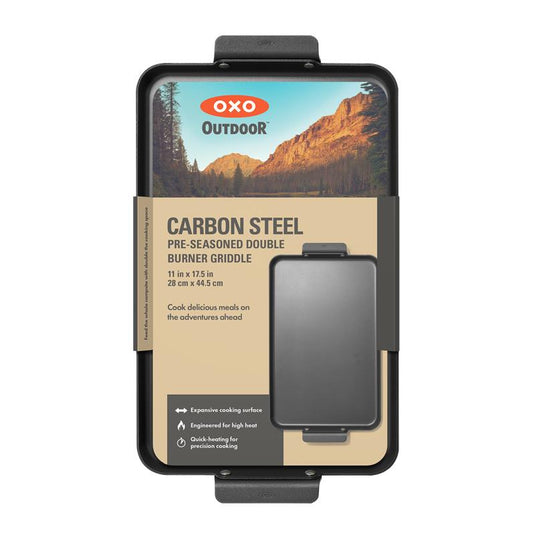 OXO Outdoor Carbon Steel Pre-Seasone Double Burner Griddle in its retail packaging, featuring a black pre-seasoned griddle insert with a tan cardboard backing and a scenic mountain graphic.