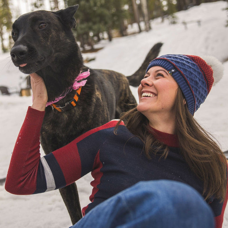 Load image into Gallery viewer, Woman in a blue and red beanie and sweater sitting in the snow with a black dog.
