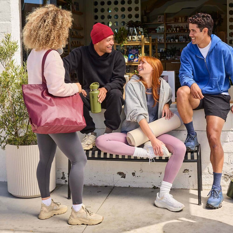 Load image into Gallery viewer, Four people in activewear pose outside a storefront. One person stands on the left carrying a large dusty rose tote bag, while three others sit and relax on a bench in the center.
