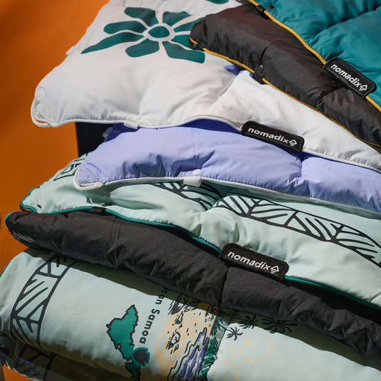 Stack of colorful sleeping bags with 'nomadix' branding on a wooden surface