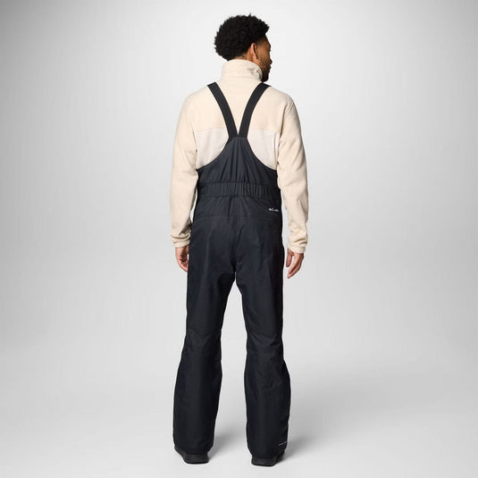 Person wearing a beige jacket and black bib overalls on a white background