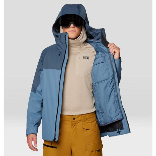 Person wearing a blue jacket with a hood, beige turtleneck, and mustard pants on a light gray background