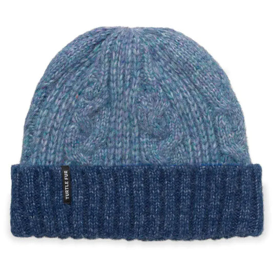 A blue and teal cable-knit beanie hat with a dark navy ribbed cuff, featuring a small black brand tag that reads 'TURTLE FUR'.